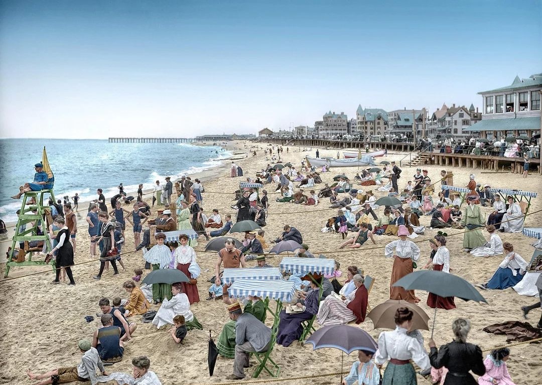 A crowded beach scene from the early 1900s shows people in vintage clothing relaxing on the sand, sitting under umbrellas, and enjoying the ocean. Lifeguards watch from a tall chair, and historic buildings line the shore in the background.