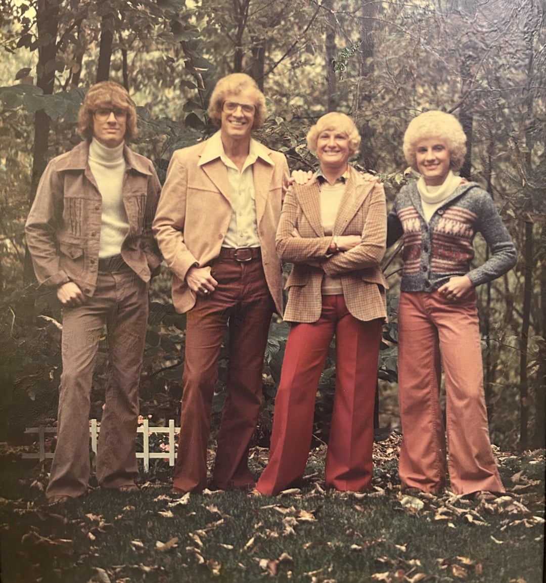 Four people stand outdoors in a wooded area wearing 1970s-style clothing, including flared pants, turtlenecks, and patterned jackets. They are smiling, with autumn leaves on the ground around them.