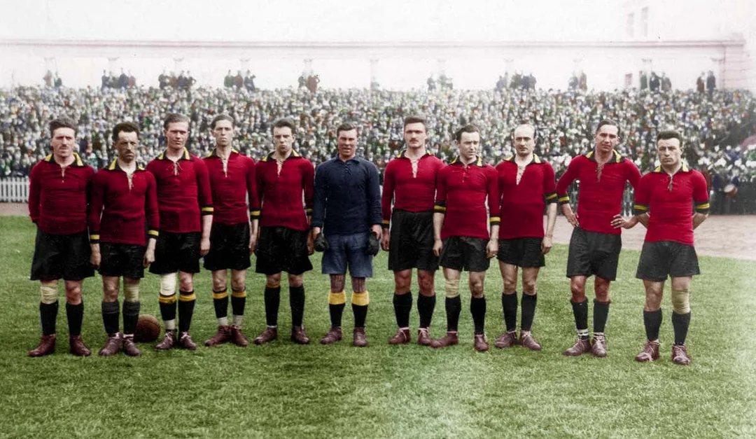 A vintage photo shows eleven male soccer players in red shirts and black shorts standing in a row on a grass field, with a crowd of spectators in the background.