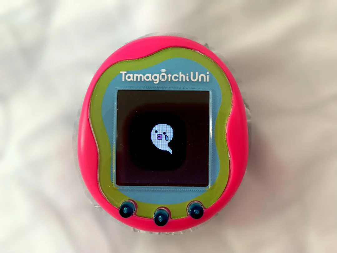 A pink and green Tamagotchi Uni device with a small screen displaying a ghost-like character on a white background.