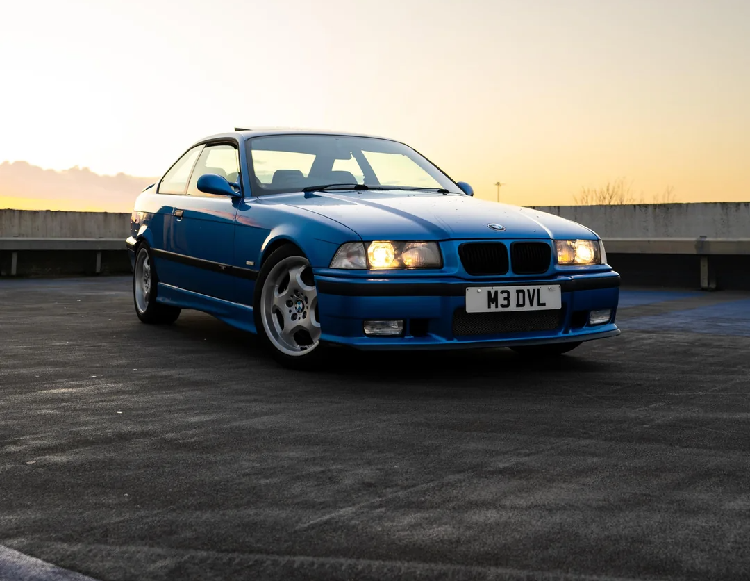 A blue BMW coupe with headlights on is parked on an empty rooftop at sunset, facing the camera at an angle. The license plate reads "M3 DVL.