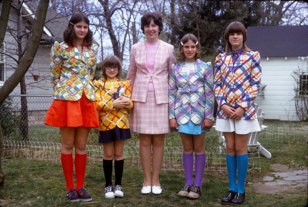 Five people stand outside in colorful, patterned outfits. Four children wear plaid jackets, skirts, and knee-high socks in various colors. An adult in the center wears a pink plaid suit. Trees and a white house are in the background.