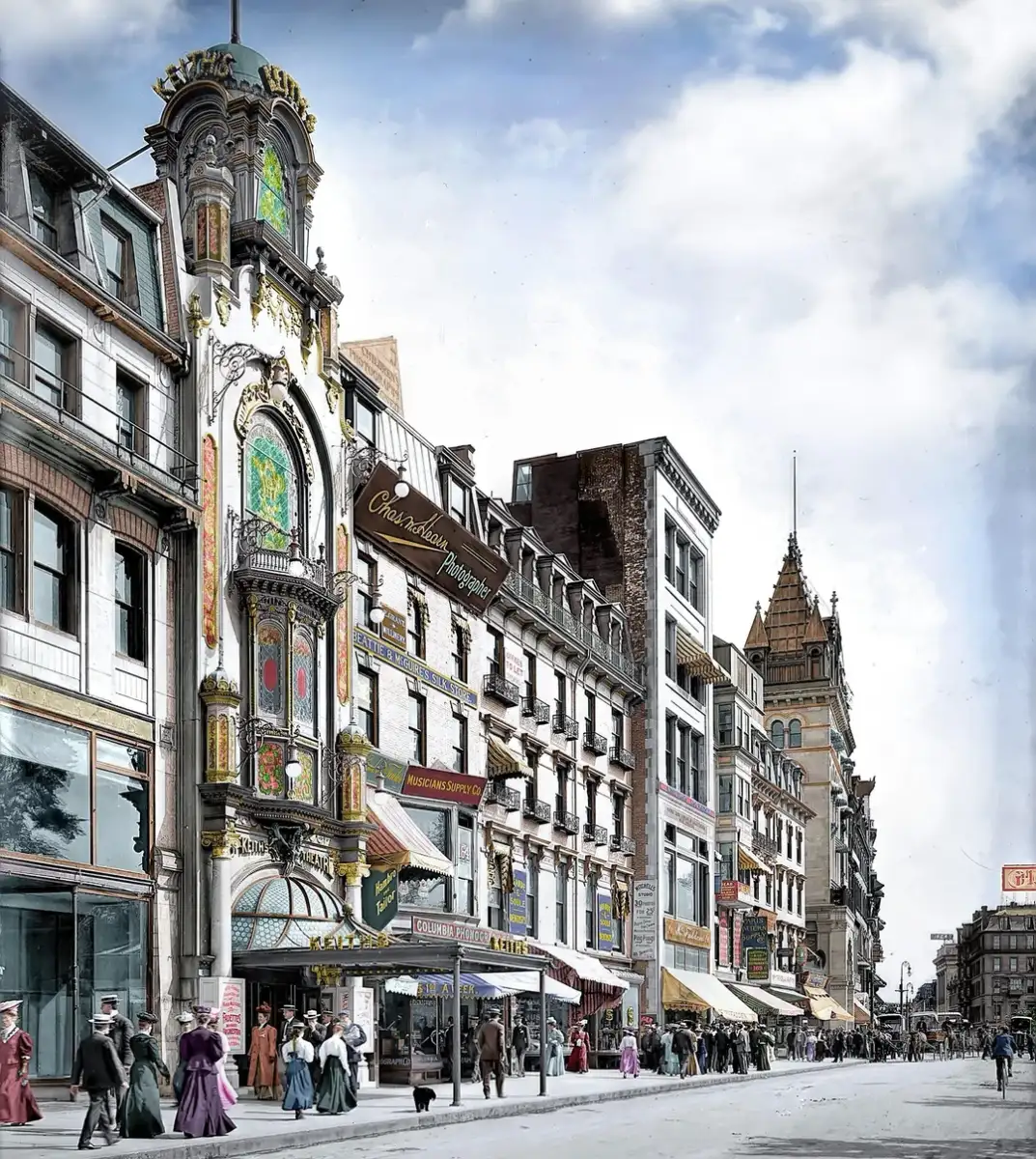 Colorized historic photo of a busy street lined with ornate buildings, including a theater with a decorative facade and stained glass. People in early 1900s attire walk and gather along the wide sidewalk.