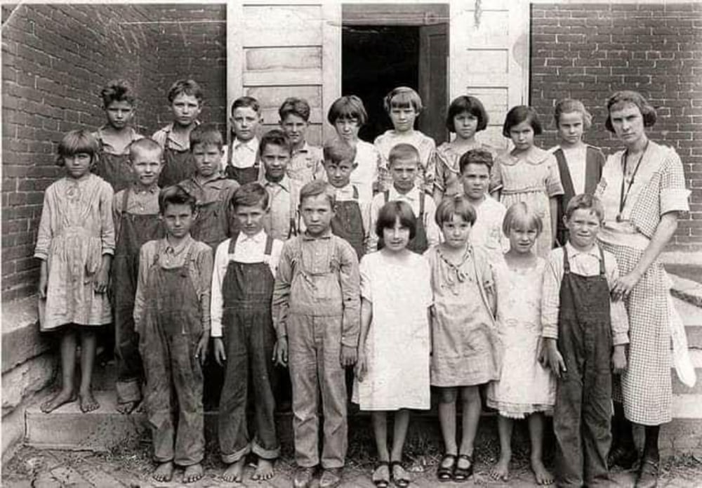 A black and white photo of a group of young children and a woman, likely their teacher, posing outside a brick building. Most children wear simple clothes; some are barefoot. The mood is serious and the image appears old-fashioned.