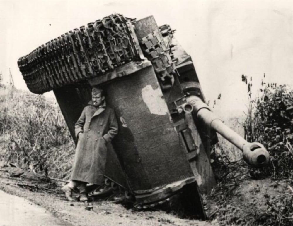 A soldier in a long coat leans against the upturned side of a large tank that has flipped onto its turret beside a road, with plants and grass in the background.