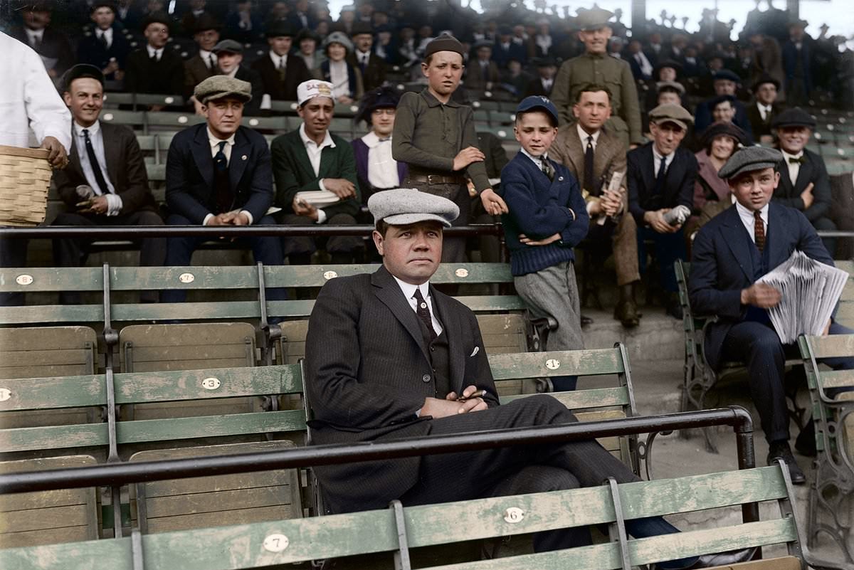 A man in a suit and flat cap sits alone in front row stadium seats, while a crowd of men, women, and boys, some in hats, watch from behind. The setting appears to be an early 20th-century baseball game.