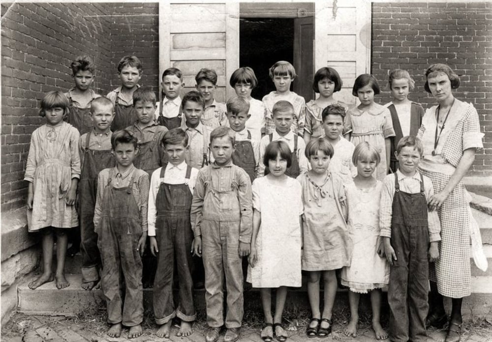 A black-and-white photo of a group of barefoot children and a female teacher standing outside a brick building, possibly a school, dressed in simple clothes, likely from the early 20th century.
