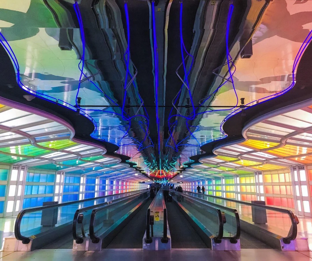 A futuristic airport walkway with moving sidewalks, illuminated by vibrant rainbow-colored lights and blue neon patterns on the ceiling, creating a tunnel-like effect with a few people in the distance.