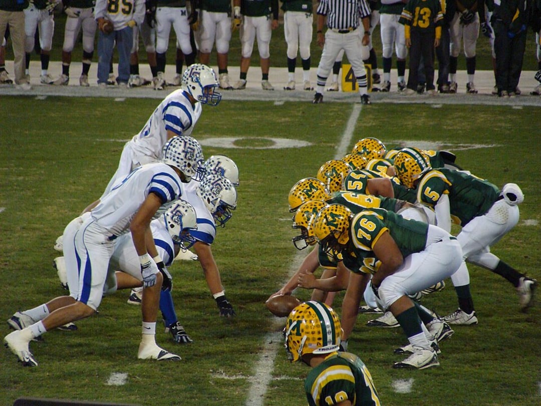 Two American football teams, one in green and yellow uniforms and the other in white and blue, line up facing each other at the line of scrimmage on a grassy field during a night game.