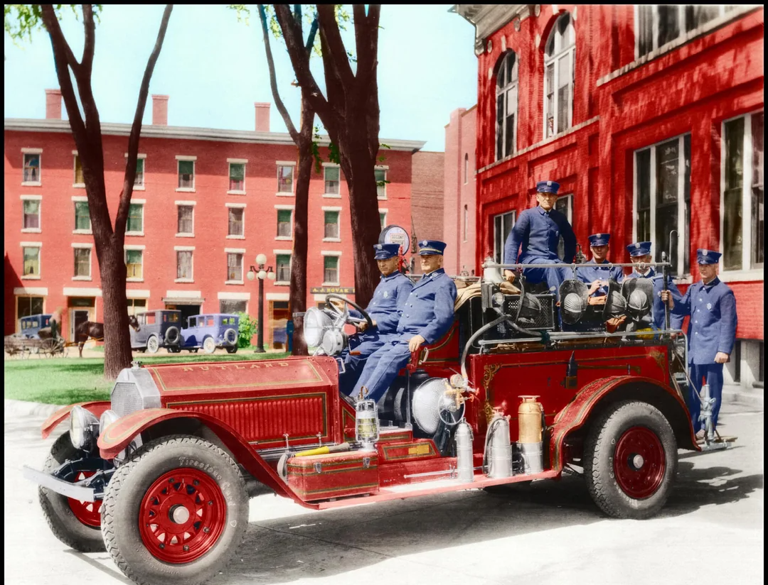 A vintage red fire truck with several uniformed firefighters is parked outside a red brick building. Other brick buildings and early 20th-century cars are visible in the background, under the shade of tall trees.