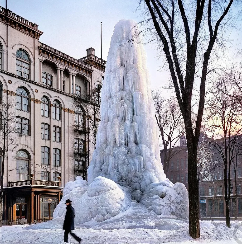 A large, icy fountain stands frozen in a snowy city square near a historic building, with a person in winter clothing walking past and bare trees framing the scene.