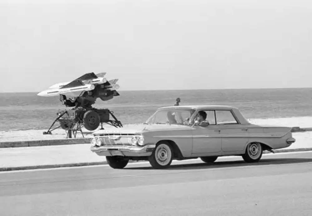 A black-and-white photo of a man driving a vintage car on a road by the beach, with a large military missile launcher positioned on the sand in the background.