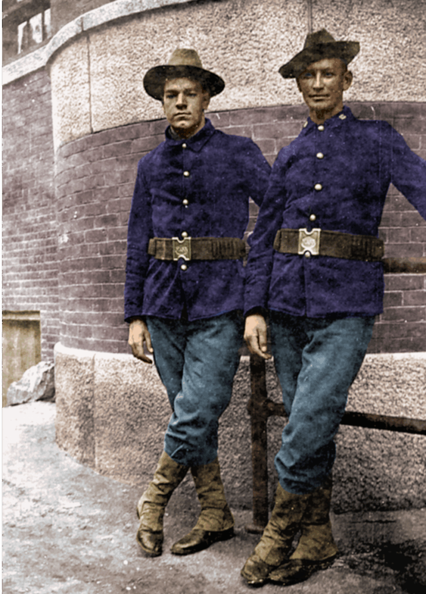Two young men in early 20th-century military uniforms with blue jackets, blue trousers, brown boots, and hats stand side by side against a brick wall, each resting an arm on a wooden rail.