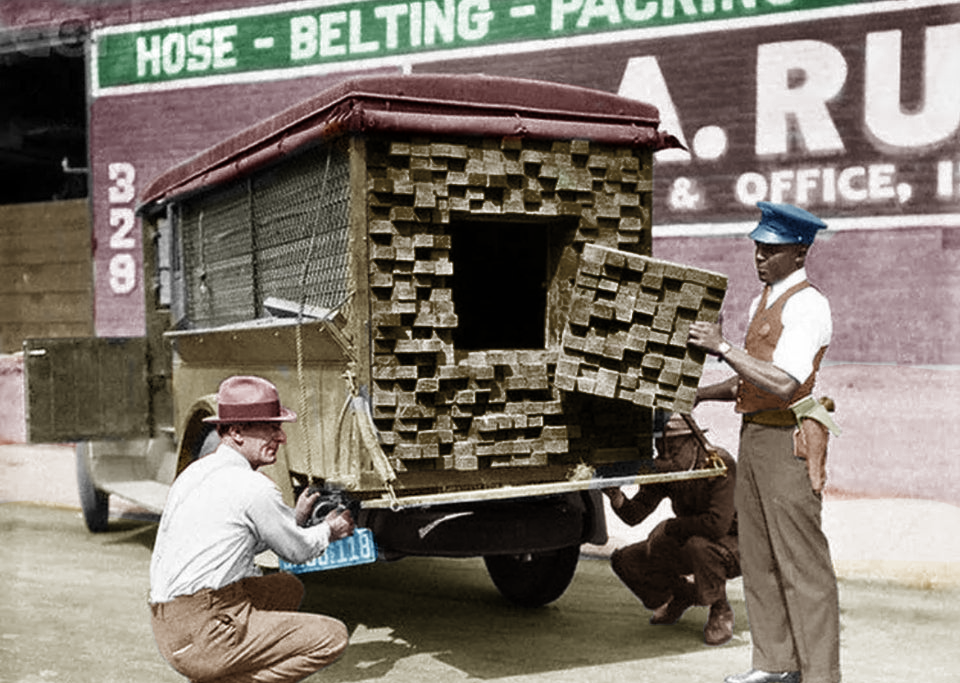 Two men show stacks of bricks hidden inside a truck with a metal cage. A police officer stands by, inspecting the secret compartment. A painted advertisement is visible on the wall behind them.