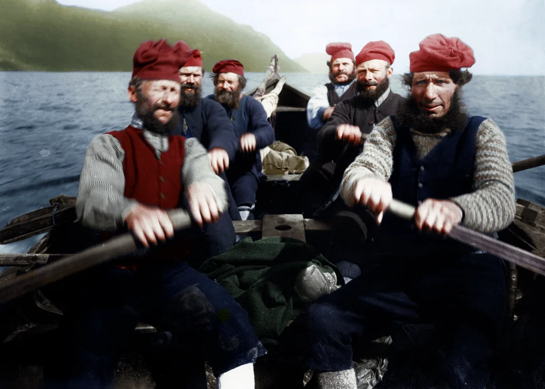 Six bearded men in red caps and traditional clothing row a wooden boat on a body of water, with green hills visible in the background under a clear sky.