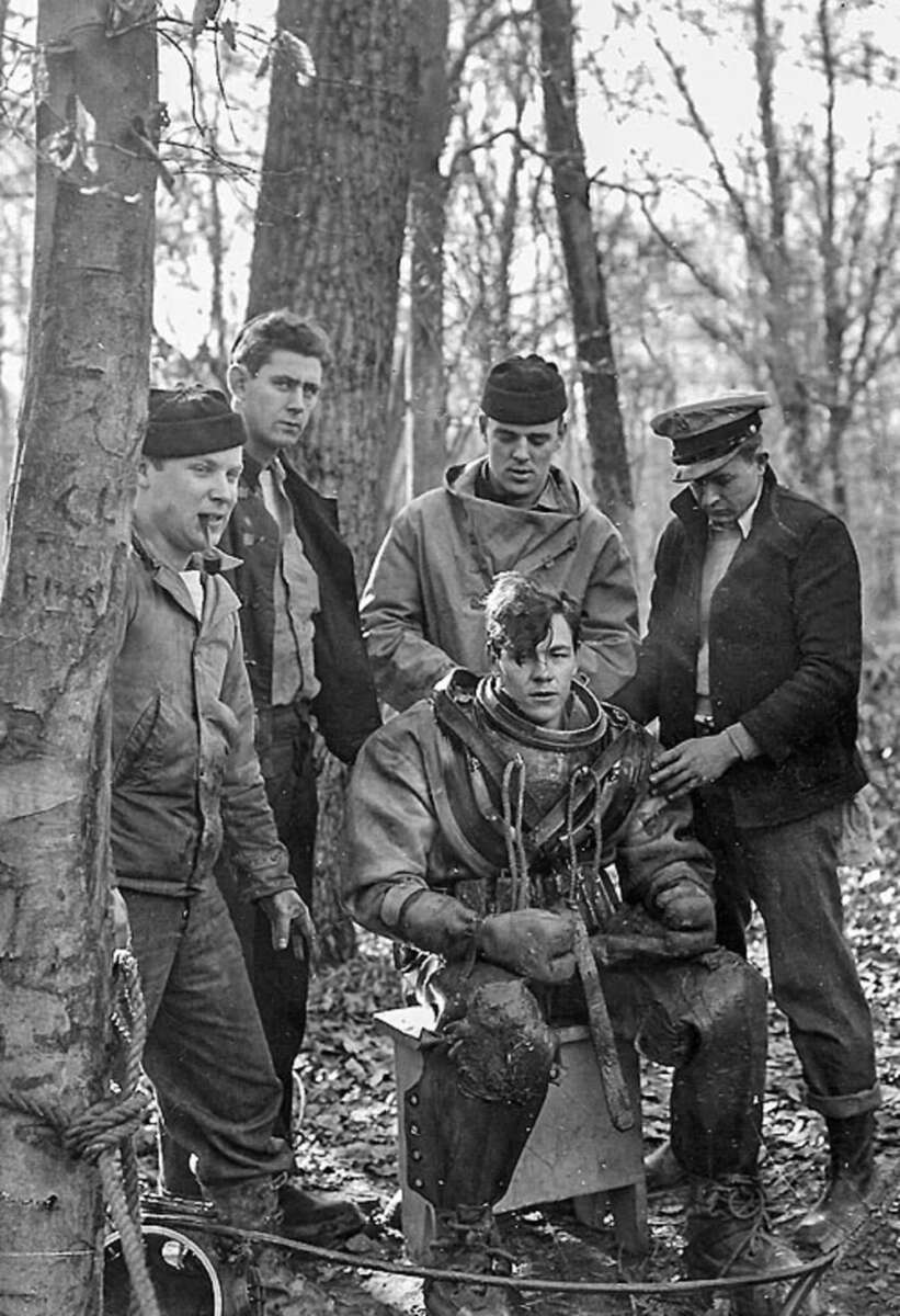 A black-and-white photo shows four men standing around a seated man wearing a vintage diving suit in a wooded outdoor setting. The standing men appear to be assisting or observing the diver.