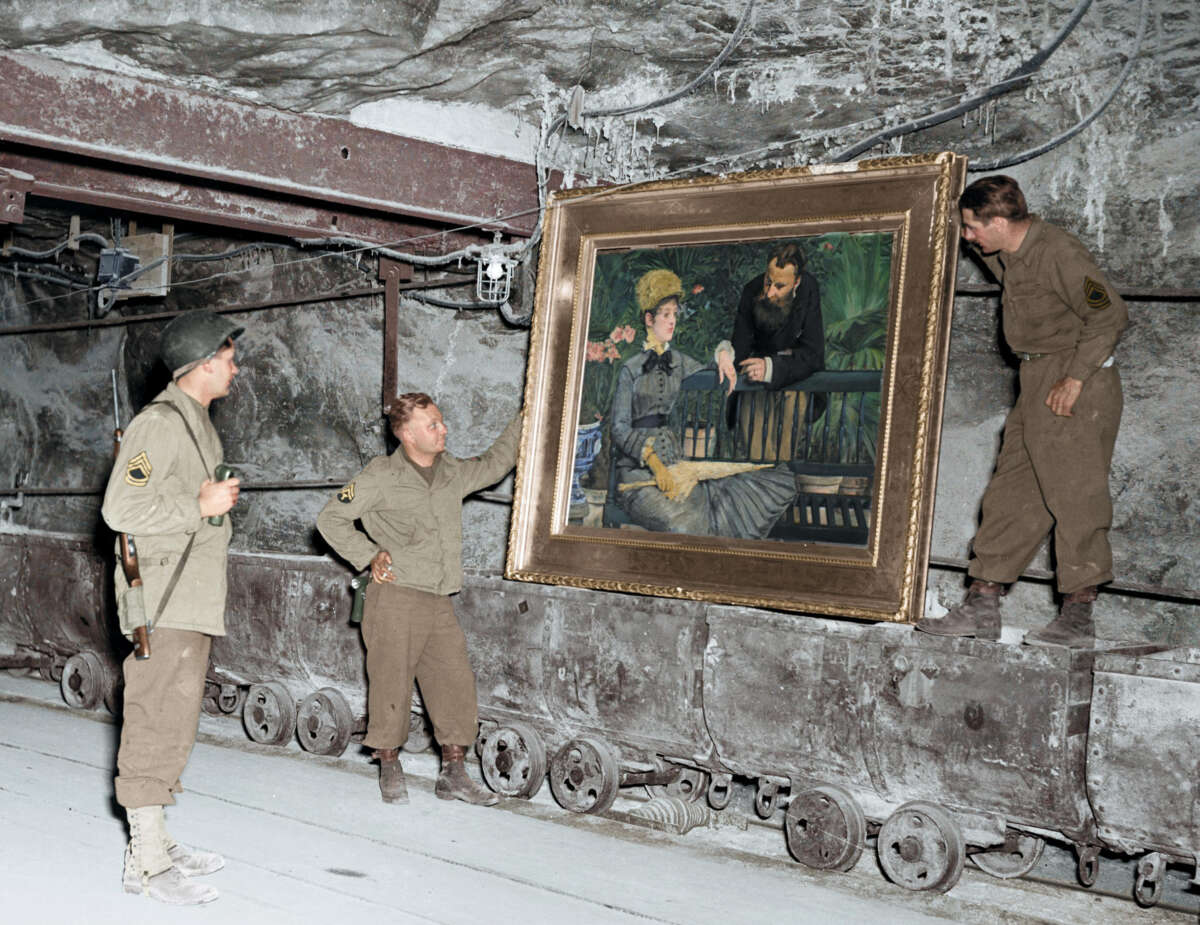 Three soldiers in uniform stand beside a large framed painting in an underground tunnel. The painting is propped on a cart, and the soldiers appear to be inspecting or recovering it. The scene is industrial and historic.