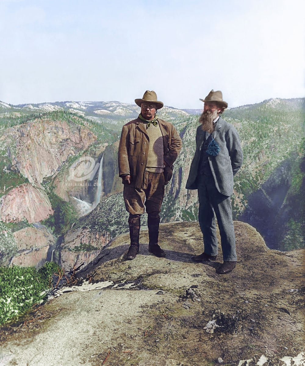 Two men in early 1900s attire stand on a rocky cliff overlooking a vast canyon and waterfall, surrounded by forested mountains under a clear sky.