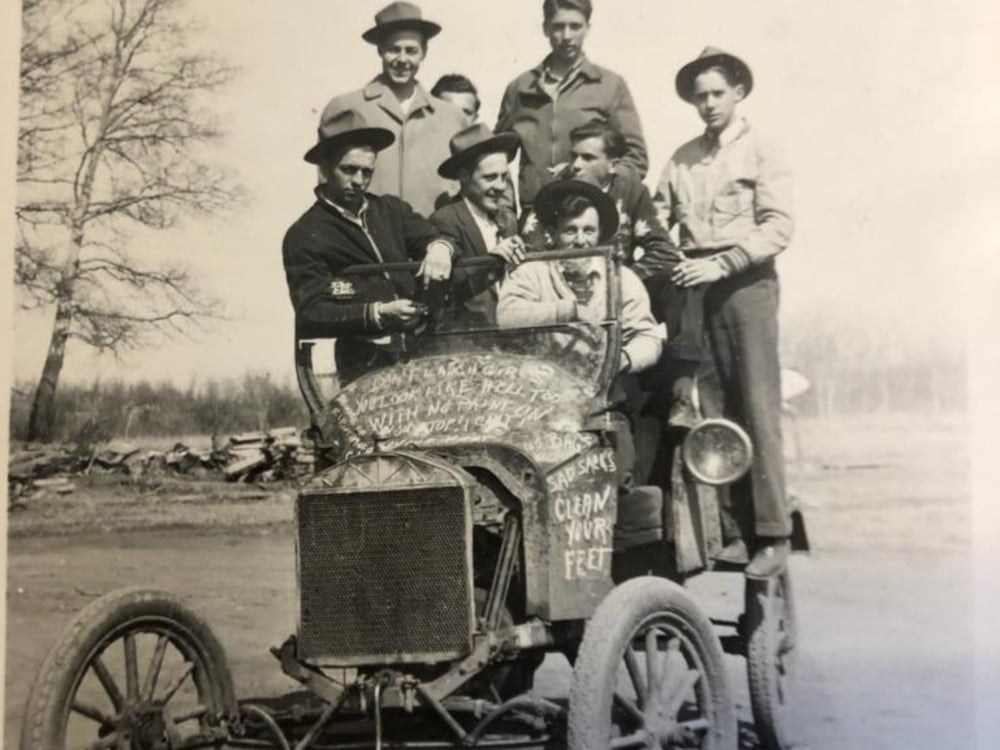 Eight men, dressed in vintage clothing, pose on and around an old-fashioned open car on a dirt road. Some sit on the vehicle, while others stand behind it; bare trees and an open field are visible in the background.