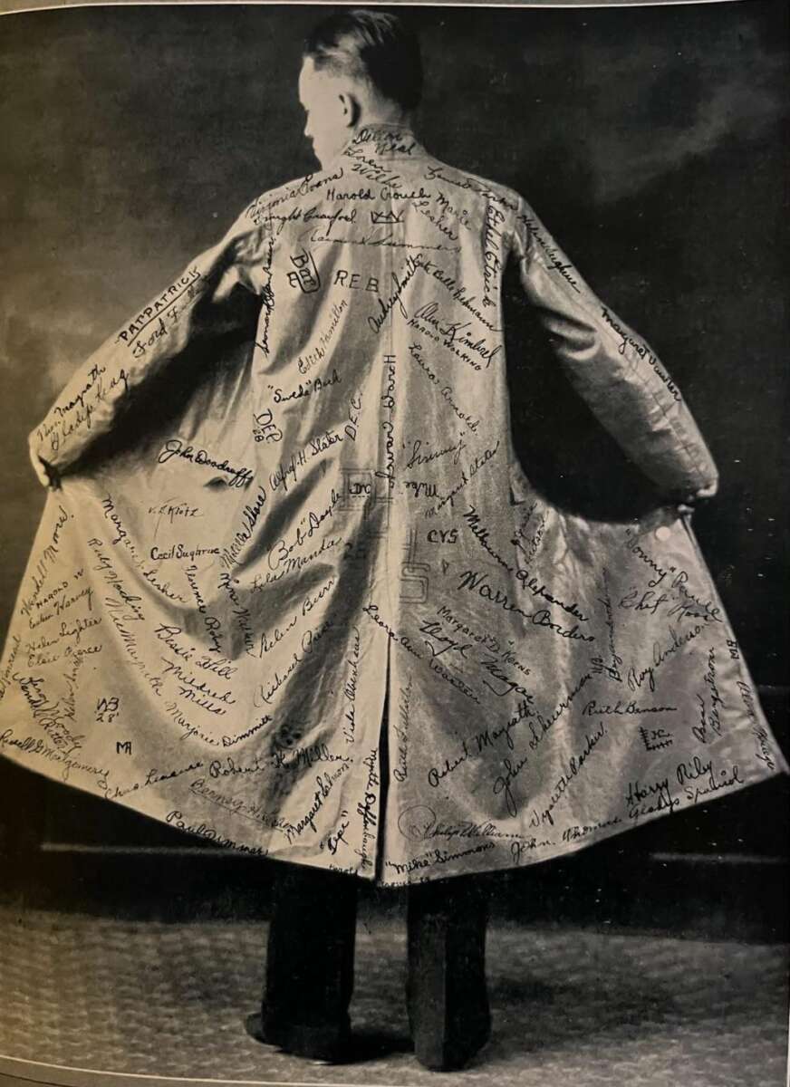 A person stands with their back to the camera, holding open a long coat covered with numerous handwritten signatures and names on the back and inner lining.