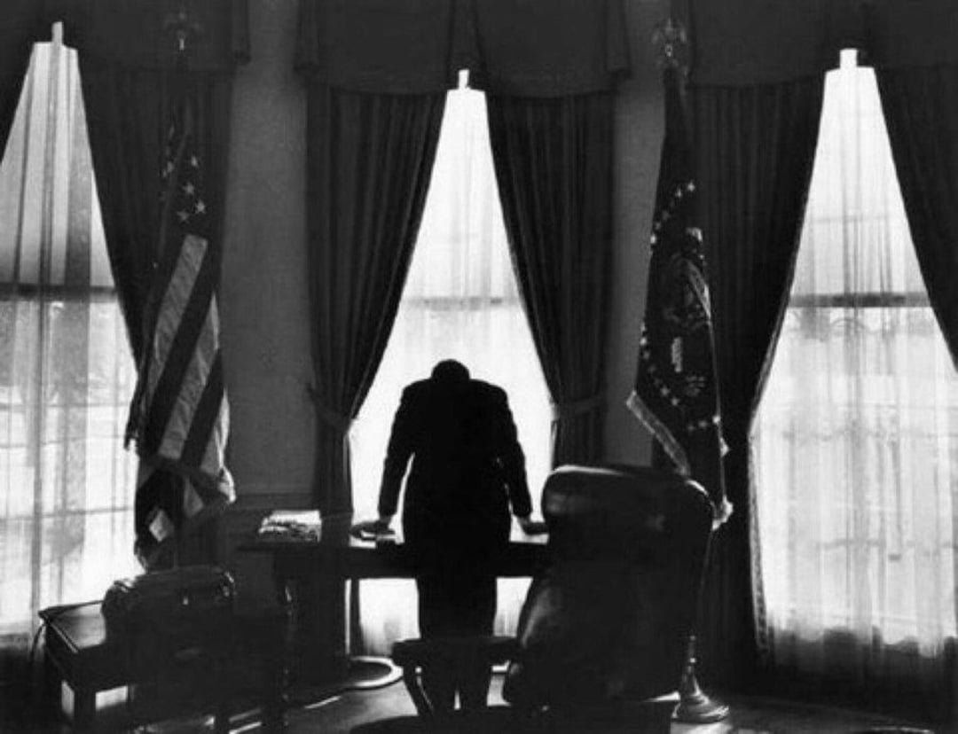 A person stands with head bowed, leaning on a desk in a dimly lit office with tall windows, flanked by the U.S. flag and presidential seal flag, silhouetted against the light.