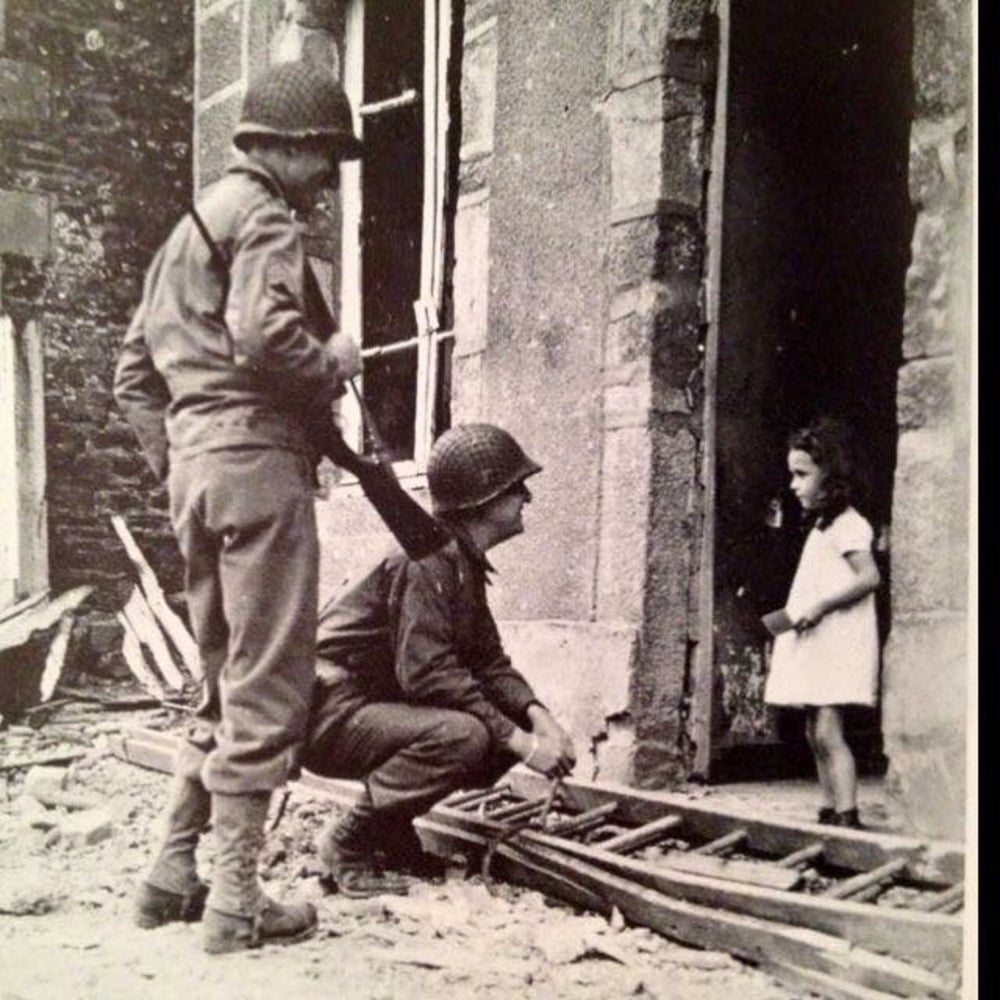 Two soldiers in uniform, one kneeling, interact with a young girl standing in a doorway of a damaged building. Debris and a ladder are on the ground, suggesting a wartime or post-conflict setting.