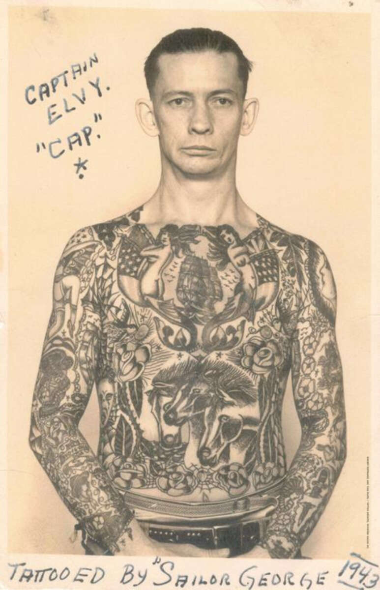 A black and white photo of a man with slicked-back hair, shirtless and heavily tattooed on his chest and arms. Handwritten text reads "CAPTAIN ELY. 'CAP.'" and "Tattooed by Sailor George 1943.