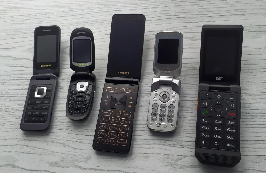 Five different flip phones from various brands are displayed open on a light gray wooden surface, showing their keypads and screens.