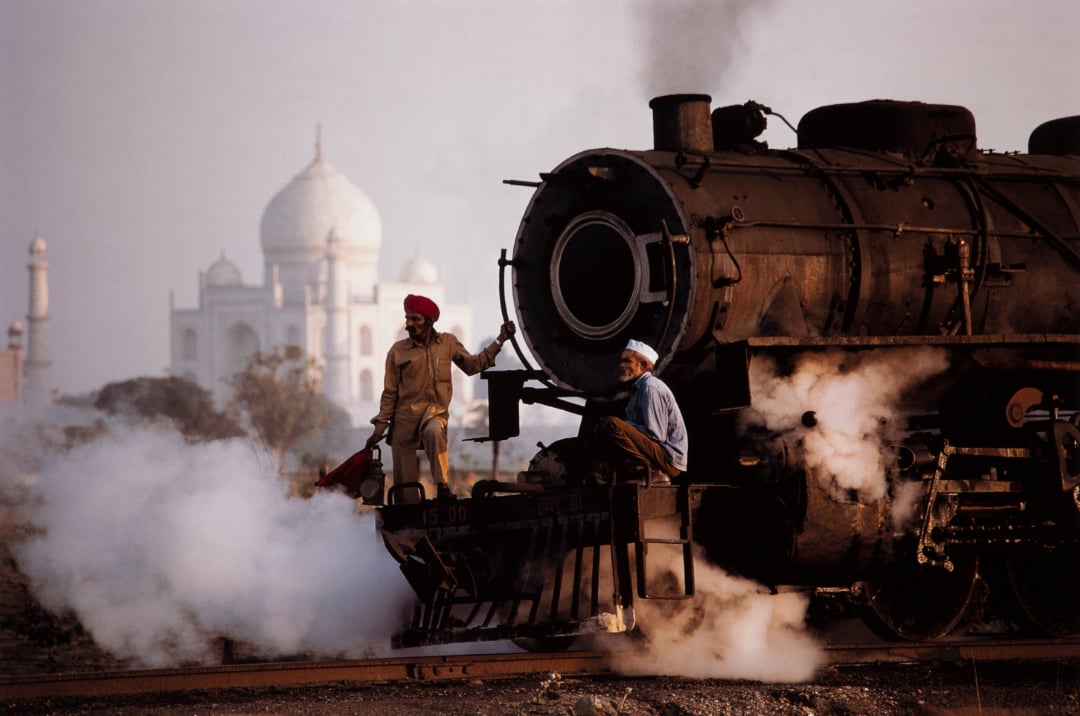 Two men stand on an old steam locomotive with white steam billowing around, while the Taj Mahal appears in the background through the haze.