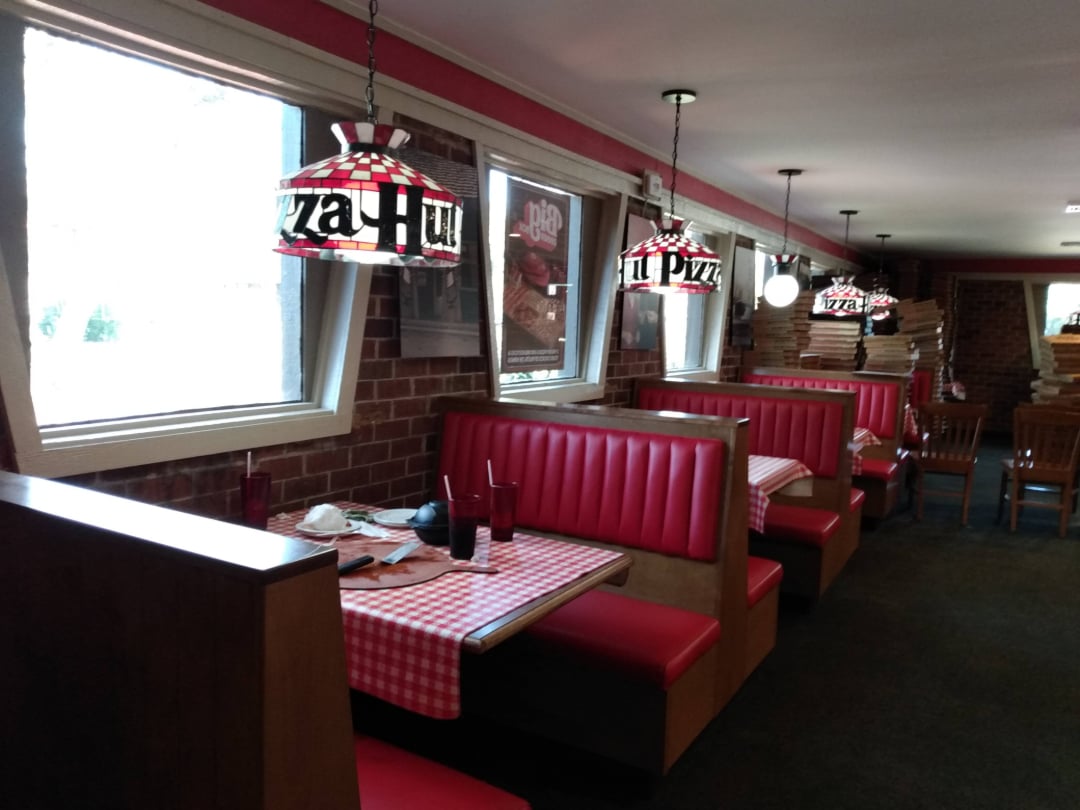 A retro-style Pizza Hut restaurant interior with red booths, checkered tablecloths, and hanging stained glass lamps. The tables are set, and sunlight streams through the windows along the brick wall.