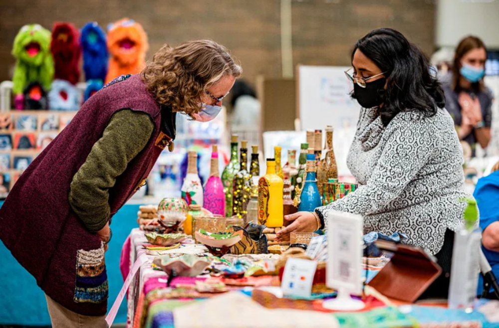 Two women wearing masks look at handmade crafts and decorated bottles on a colorful table at an indoor market or craft fair. Other booths and people can be seen in the background.
