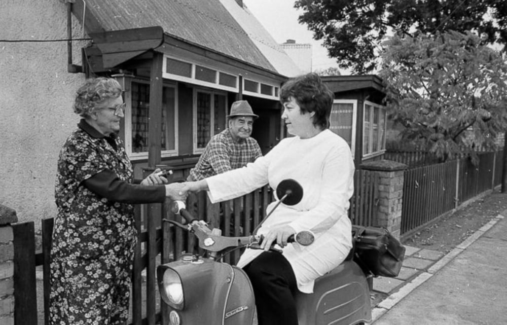 A woman on a scooter shakes hands with an older woman standing at a gate, while an older man watches from behind the fence in front of a house. The scene appears friendly and takes place on a residential street.
