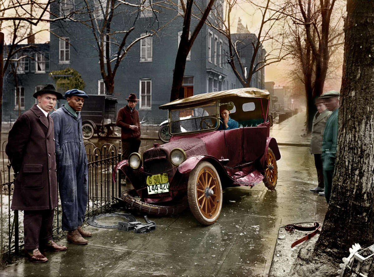 A vintage car with a damaged front wheel sits on a wet city street. Four men stand nearby, one in coveralls, while another man sits inside the car. Leafless trees and old buildings line the background.