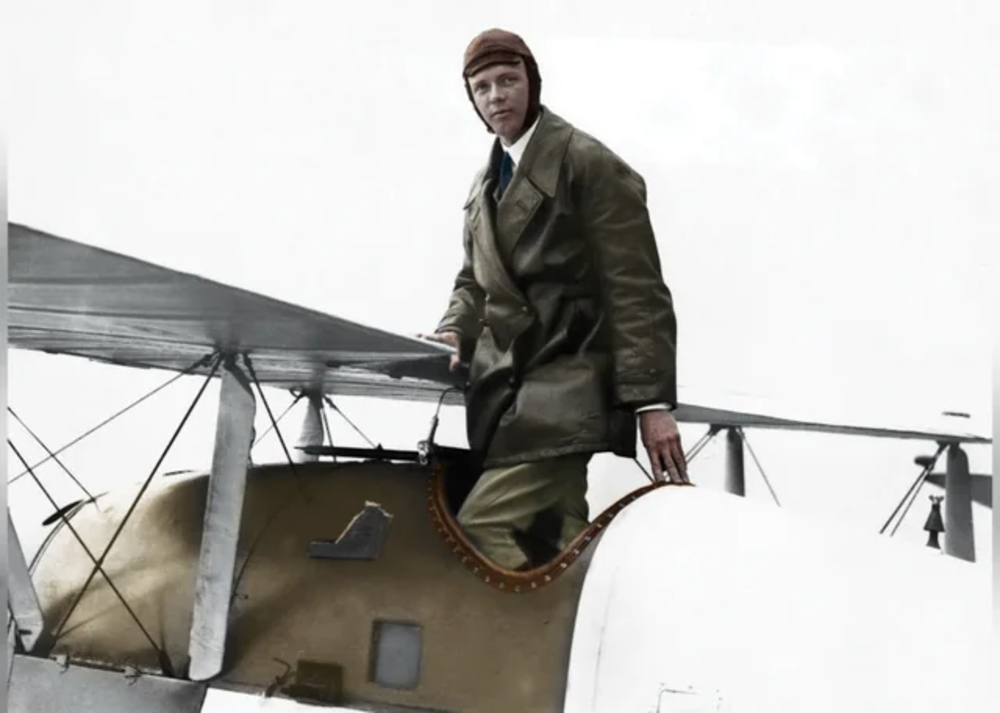 A man in a leather aviator cap and coat stands on the wing of a vintage biplane, holding onto the cockpit. The image appears to be taken in early aviation history, with a muted and overcast sky in the background.
