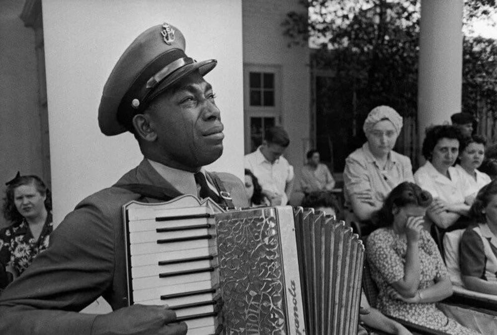 A uniformed man plays an accordion outdoors, looking upwards. Several women and another man stand or sit behind him, some watching, some looking away or appearing pensive.