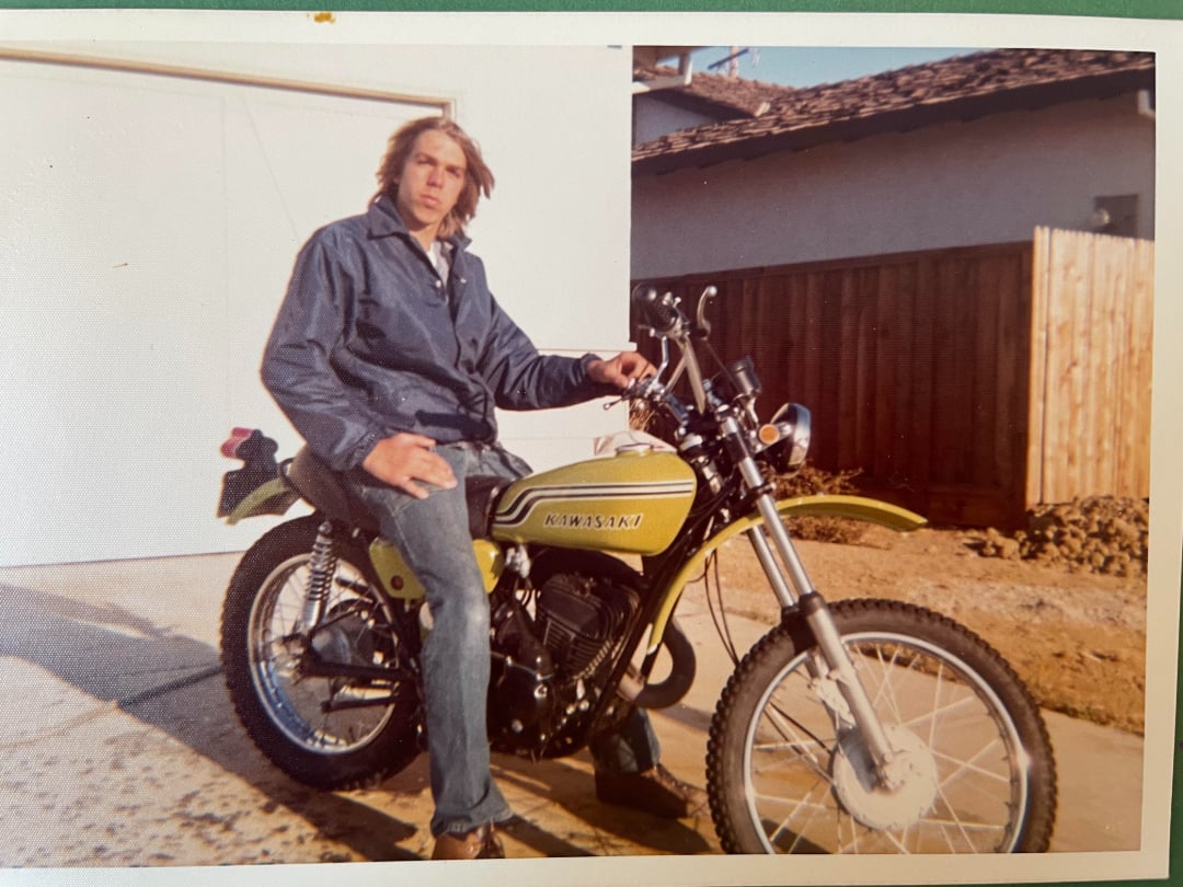 A young man in a dark jacket and jeans sits on a yellow Kawasaki motorcycle parked in a driveway, with a wooden fence and white garage in the background.