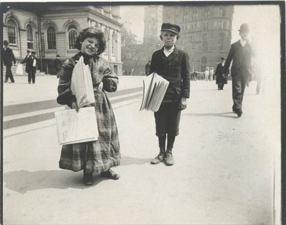 Two children stand on a city sidewalk holding newspapers. The girl smiles at the camera, wearing a plaid dress, while the boy stands nearby in a cap and suit. People and historic buildings are visible in the background.