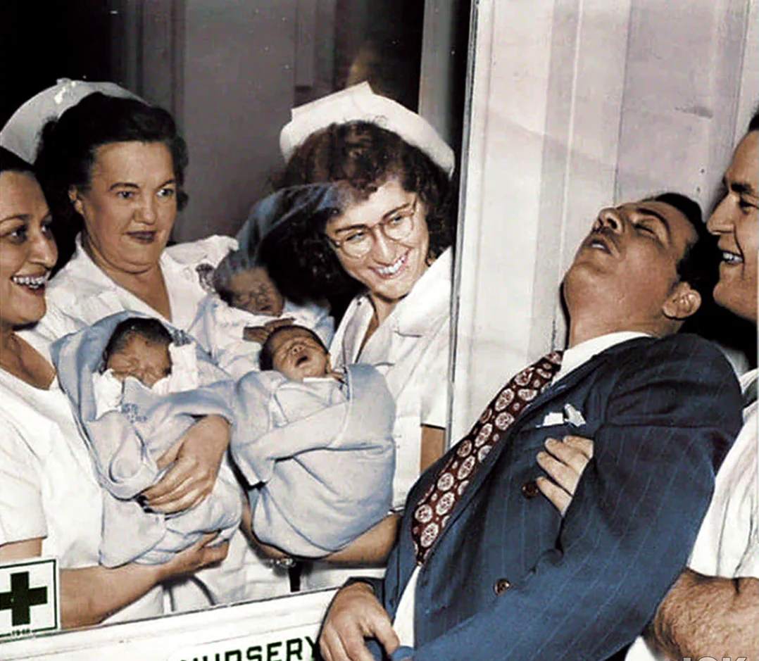 Three nurses holding newborn babies smile at a glass window, while two suited men stand outside; one man appears to have fainted or is overwhelmed with emotion, leaning back with his eyes closed.