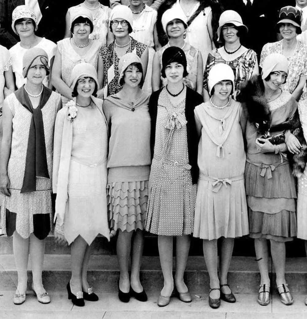 A group of women from the 1920s pose together on stairs, wearing cloche hats, dresses with dropped waists, and long necklaces. Most are smiling, and their flapper-style outfits reflect the fashion of the era.