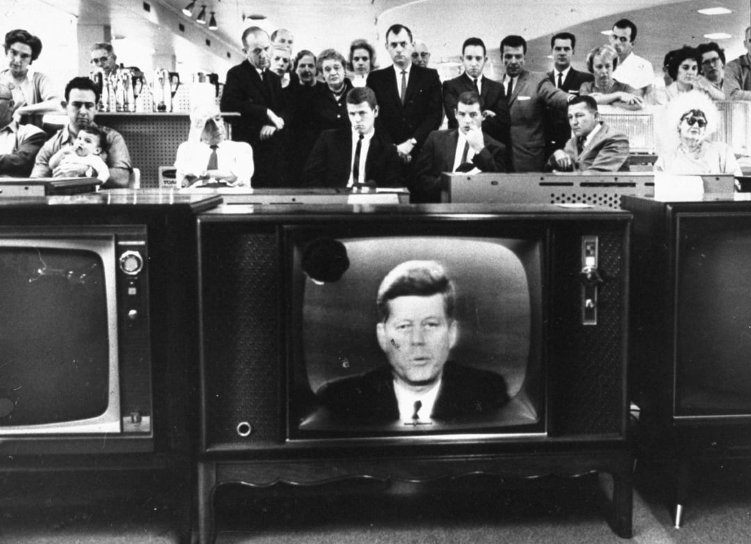 People gather in a store, watching President John F. Kennedy deliver a televised speech on several TVs. Kennedy’s face appears on the central screen, while the audience looks on with serious expressions.