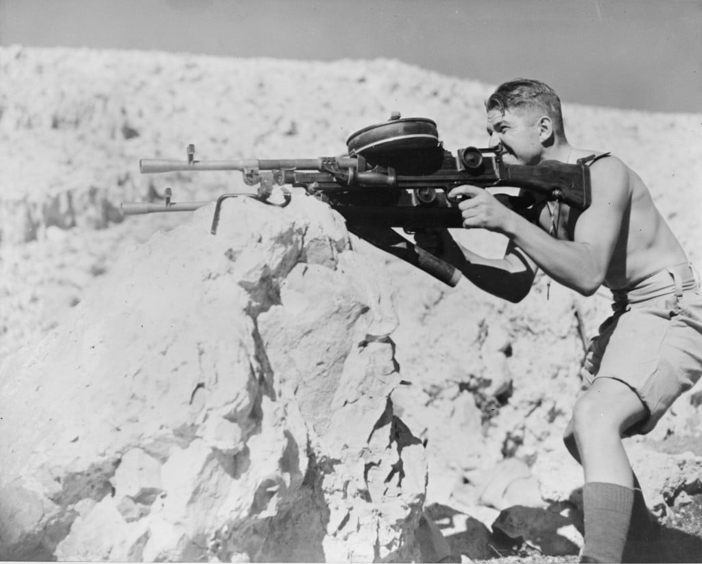 A soldier in shorts aims a large machine gun from behind a rocky outcrop in a desert landscape, under a clear sky.