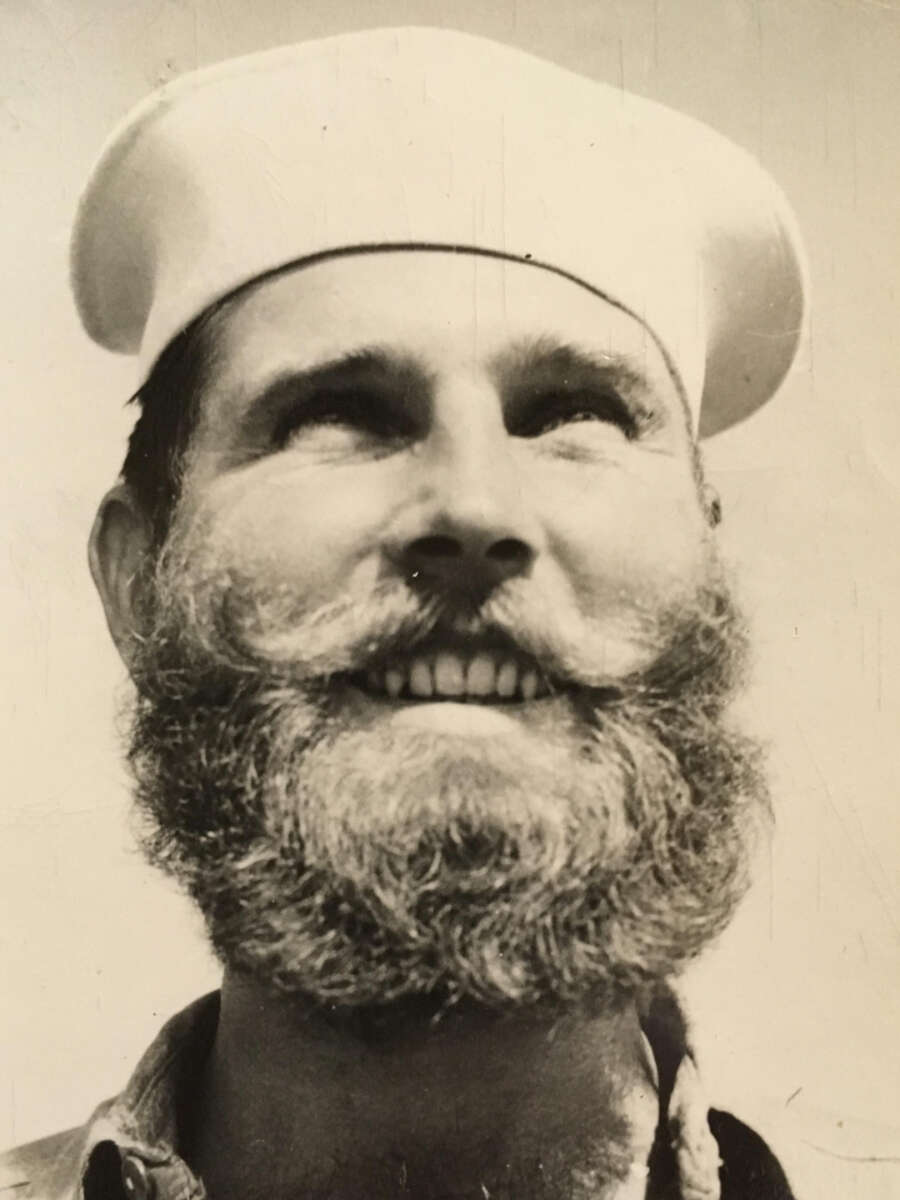 A bearded man wearing a white sailor hat smiles and looks upward, pictured in black and white with sunlight illuminating his face.