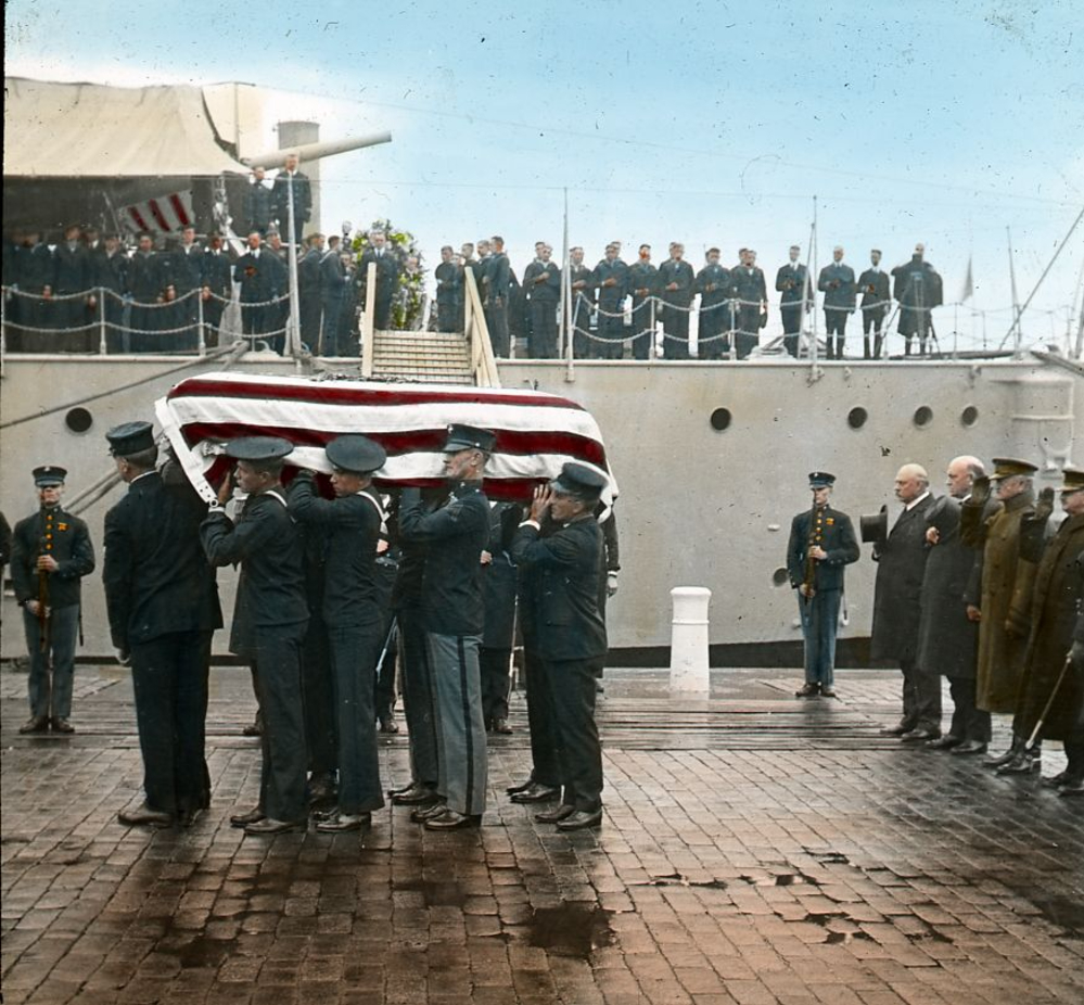 Pallbearers in military uniforms carry a flag-draped coffin near a ship, while uniformed personnel and civilians stand in the background on a rainy day.