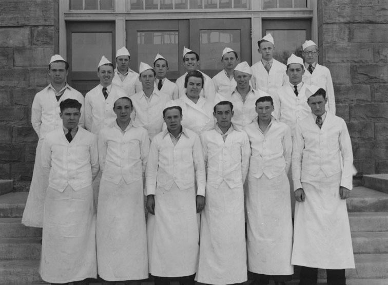 A group of seventeen men wearing white uniforms and hats stand in three rows on steps outside a stone building with large windows, posing for a formal group photo.