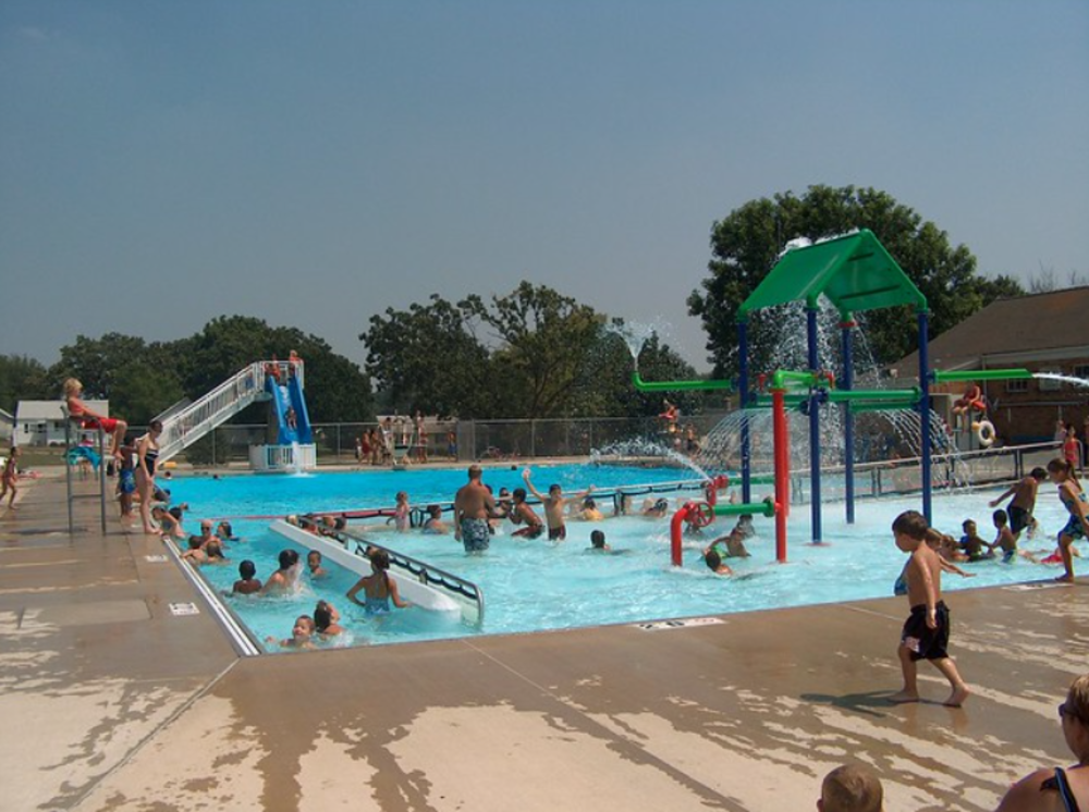 Children and adults enjoy a busy outdoor public pool with a waterslide, lifeguard, and water play structures on a sunny day. Some people are swimming while others play or walk around the wet poolside.