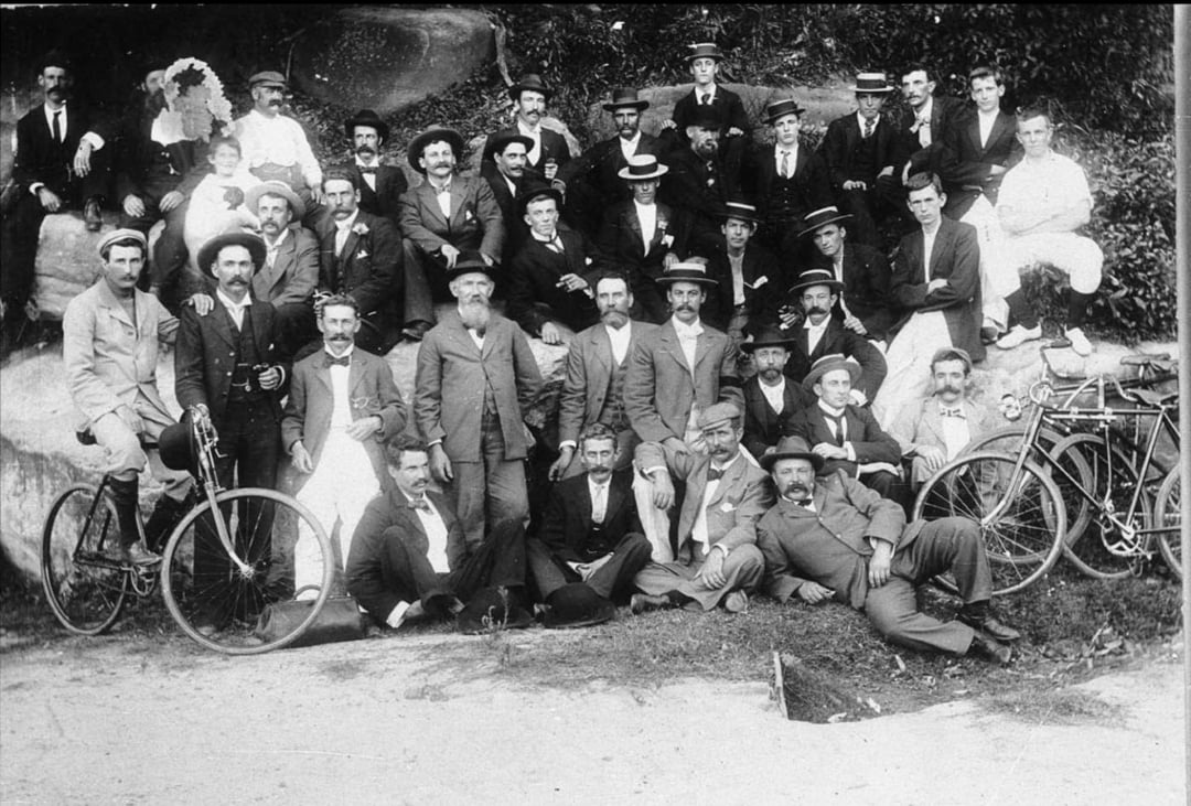 A large group of men in suits and hats pose outdoors with bicycles on a grassy slope, some standing and others sitting or reclining, in a black-and-white vintage photograph from the early 20th century.