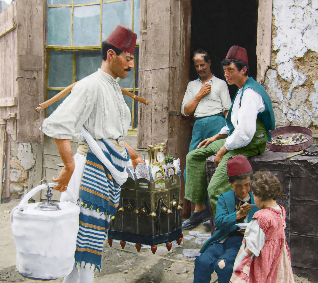 A man in traditional clothing and a fez serves drinks from a dispenser to children and adults seated outside a weathered building. The group appears relaxed and engaged in conversation.