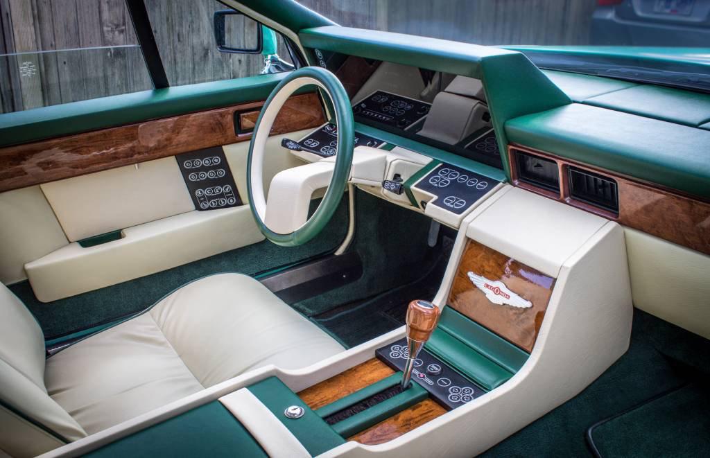 Interior of a vintage car featuring green and cream leather seats, wood trim, a unique rectangular steering wheel, and a central console with numerous buttons and a wooden gear shifter.