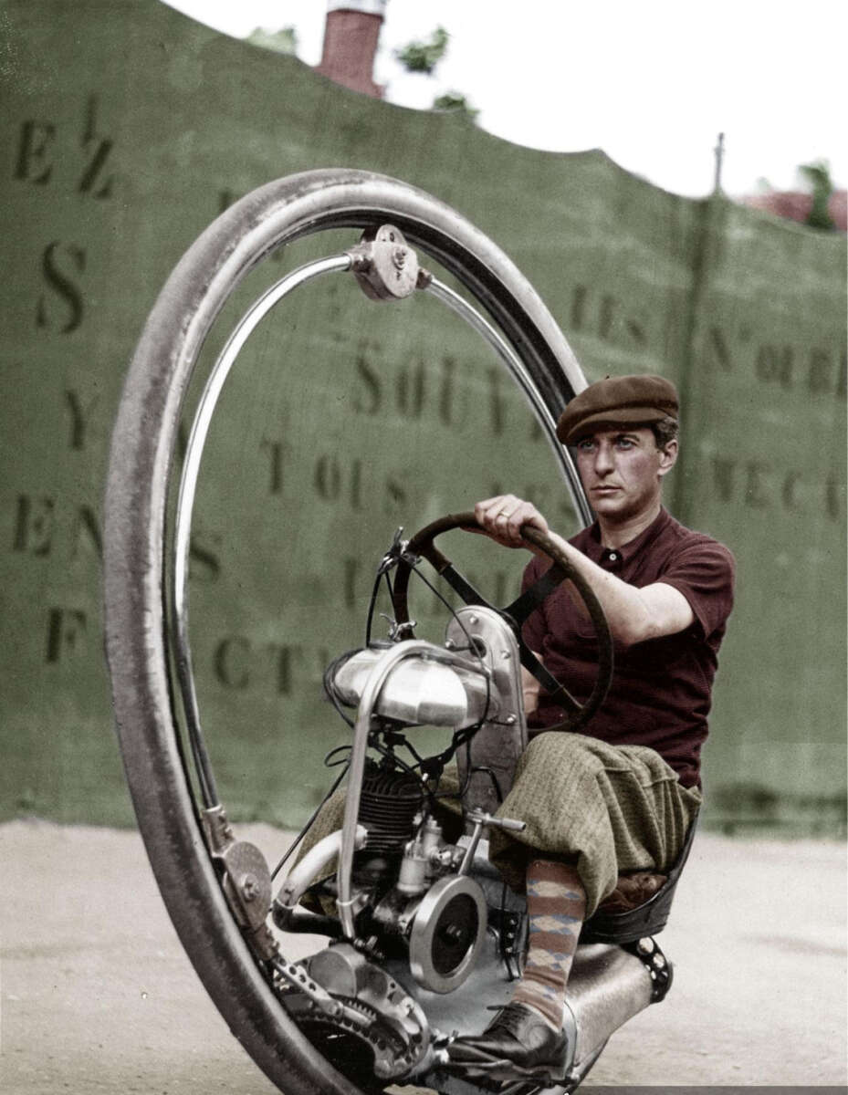 A man wearing a cap and patterned socks sits and steers a vintage monowheel motorcycle, with a large single wheel surrounding him, in front of a green wall with faded writing.