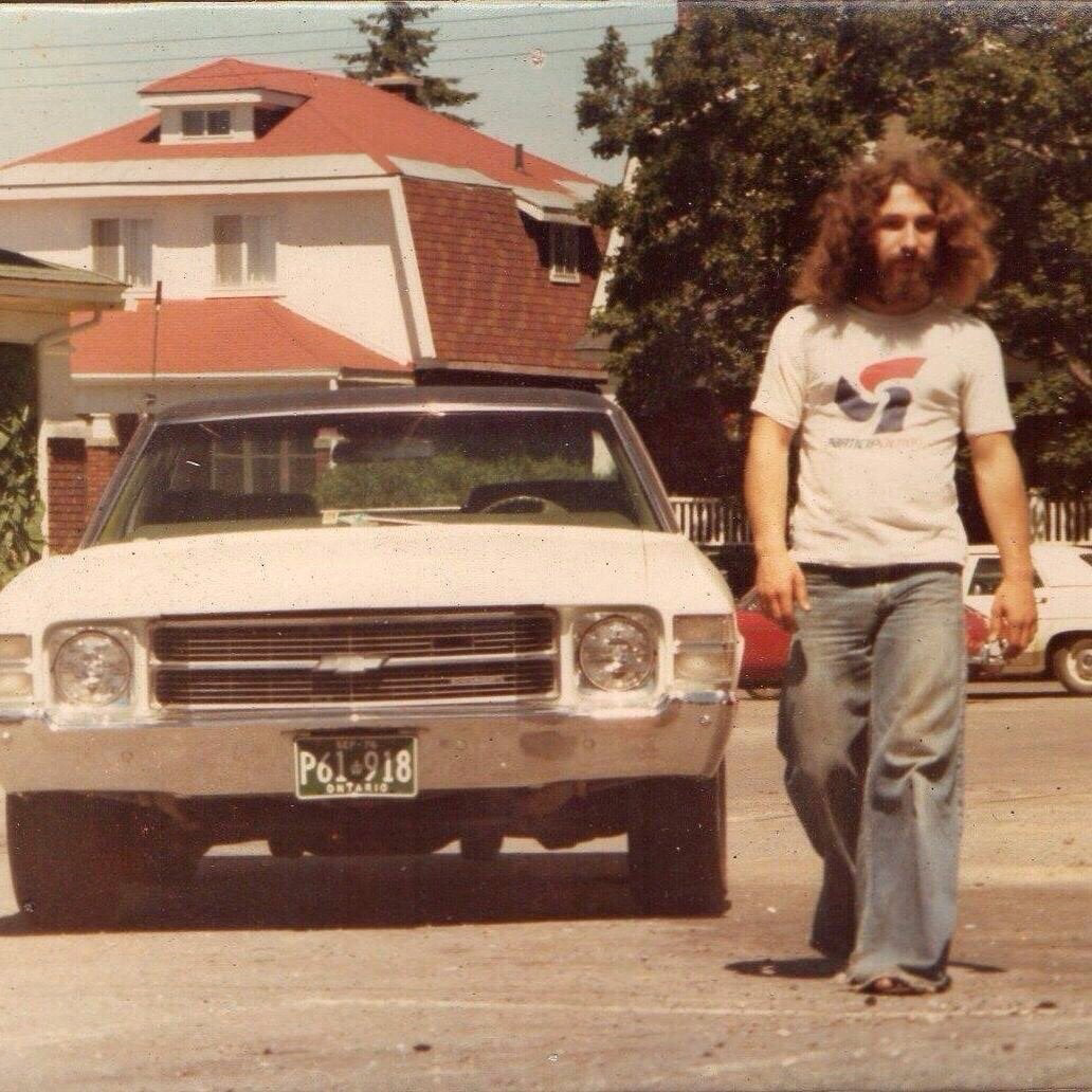 A man with long curly hair and a beard, wearing a white t-shirt and jeans, walks in a parking lot beside a vintage white car with a green license plate. A house and trees are visible in the background.